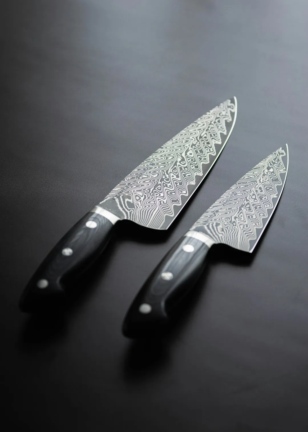 Two kitchen knives of different lengths with dark handles and patterned blades on a dark surface.