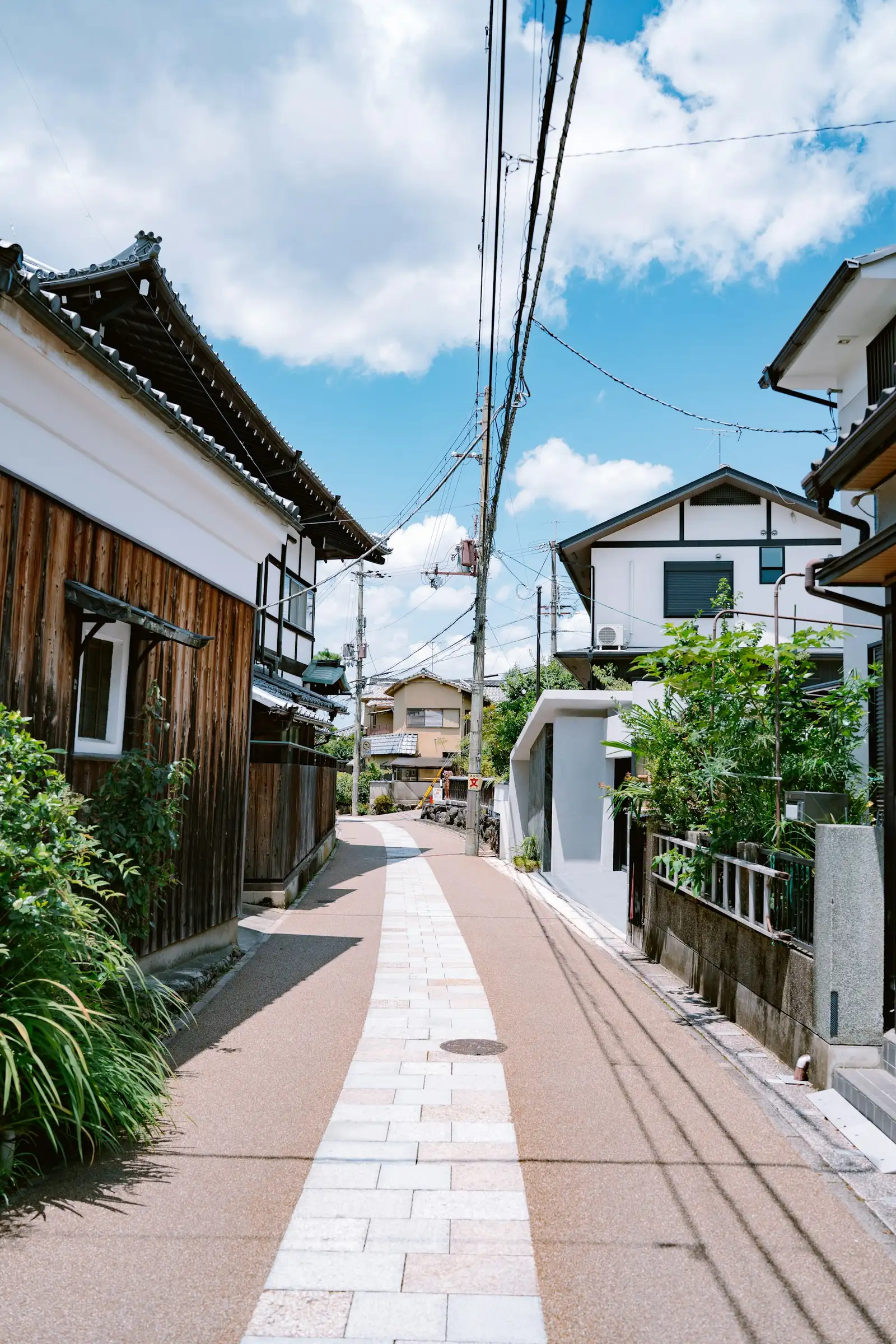 A narrow residential street in Japan lined with wooden and stucco houses, overhead power lines, under a blue sky.