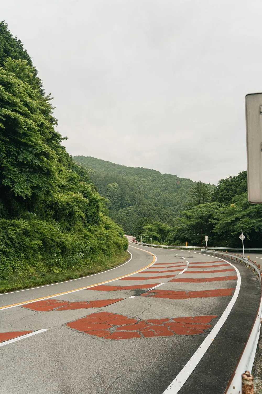A curving road through wooded hills in Japan with red-painted lane markings and a guardrail.