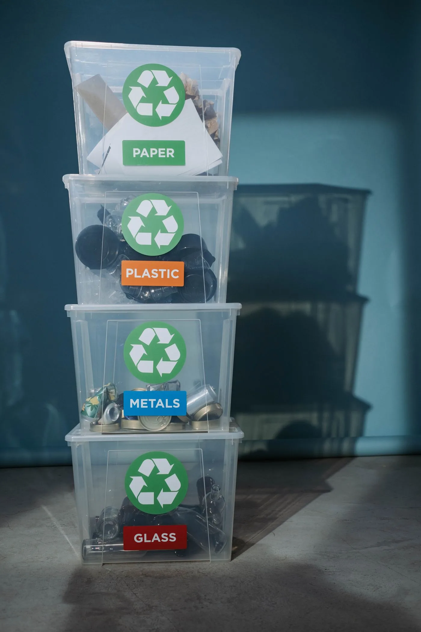 Four stacked clear storage boxes labeled paper, plastic, metals and glass, each holding the matching material.