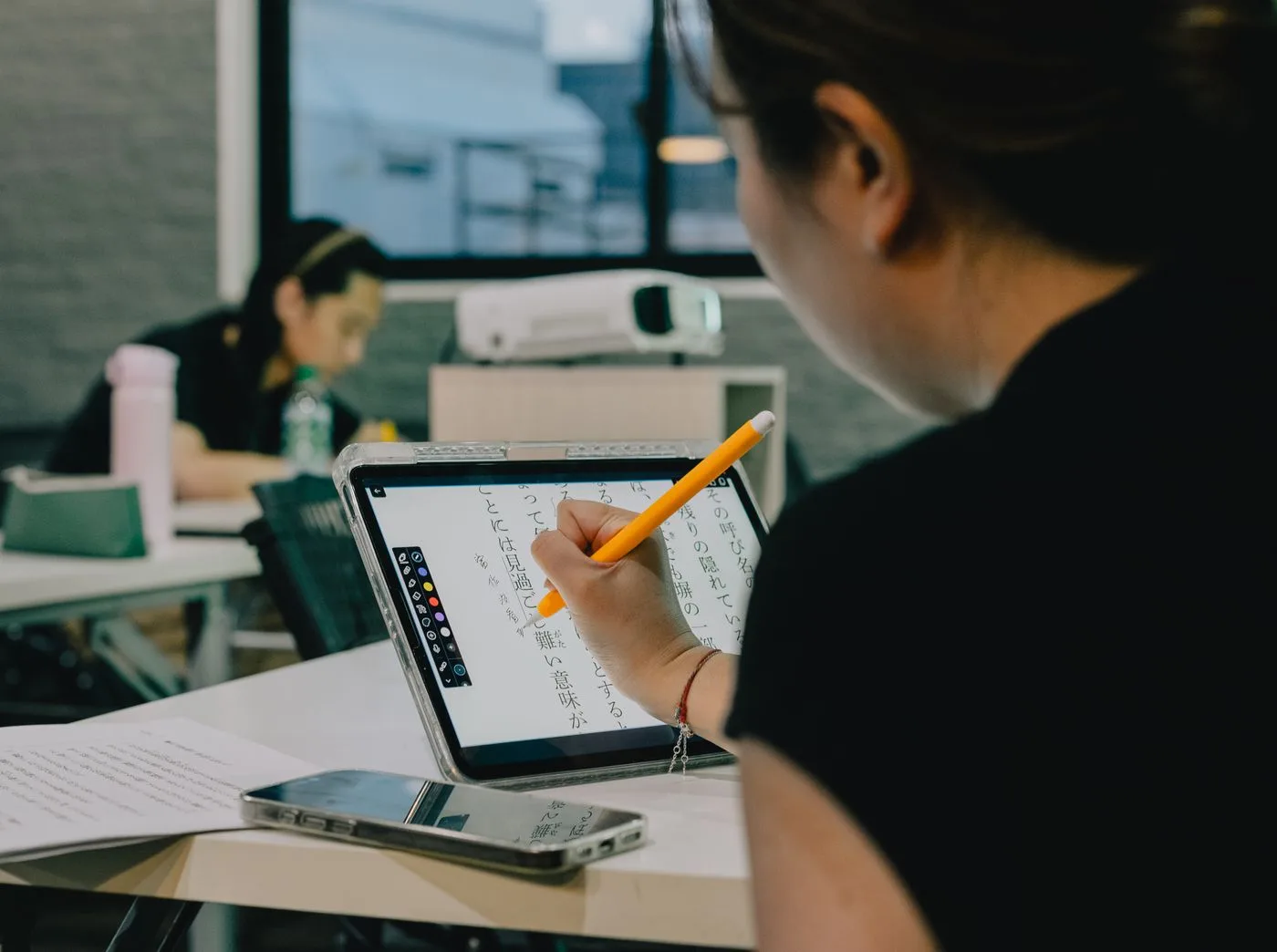 A person in a classroom writing on a tablet that displays vertical Japanese text with reading aids alongside the characters.