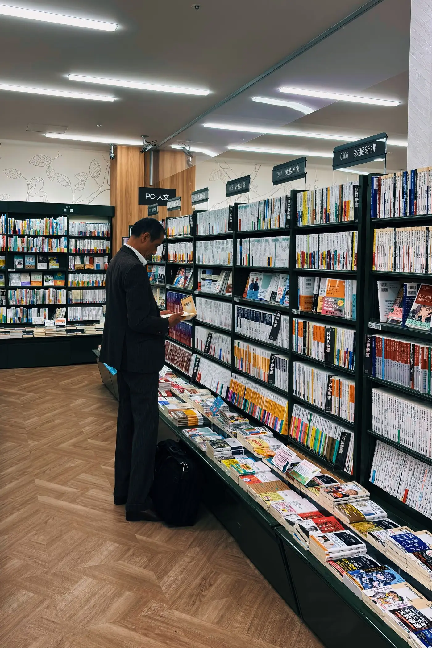 A modern Japanese bookshop aisle, with a man in a suit reading a paperback beside shelves of Japanese books.