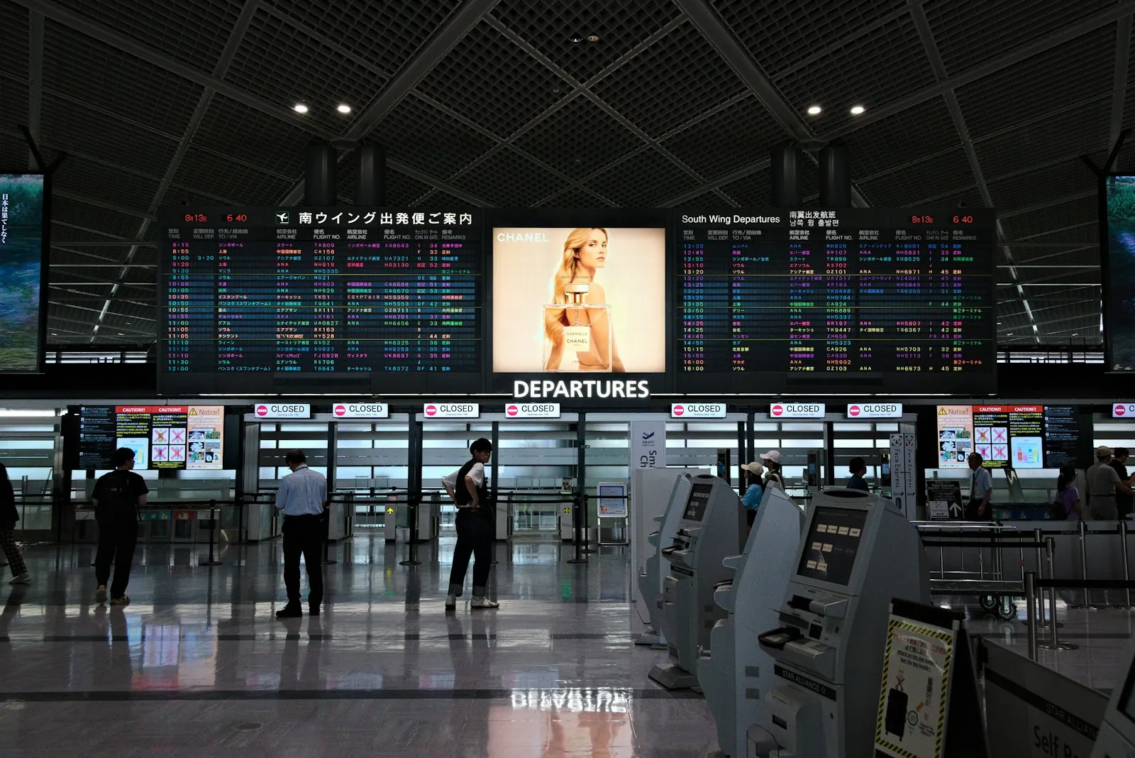 A departures hall at a Japanese airport with self-service check-in kiosks and flight information boards.