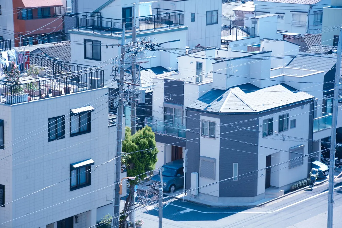 Aerial view of a dense Japanese residential neighborhood with tiled and metal rooftops, balconies, utility poles and overhead power lines.