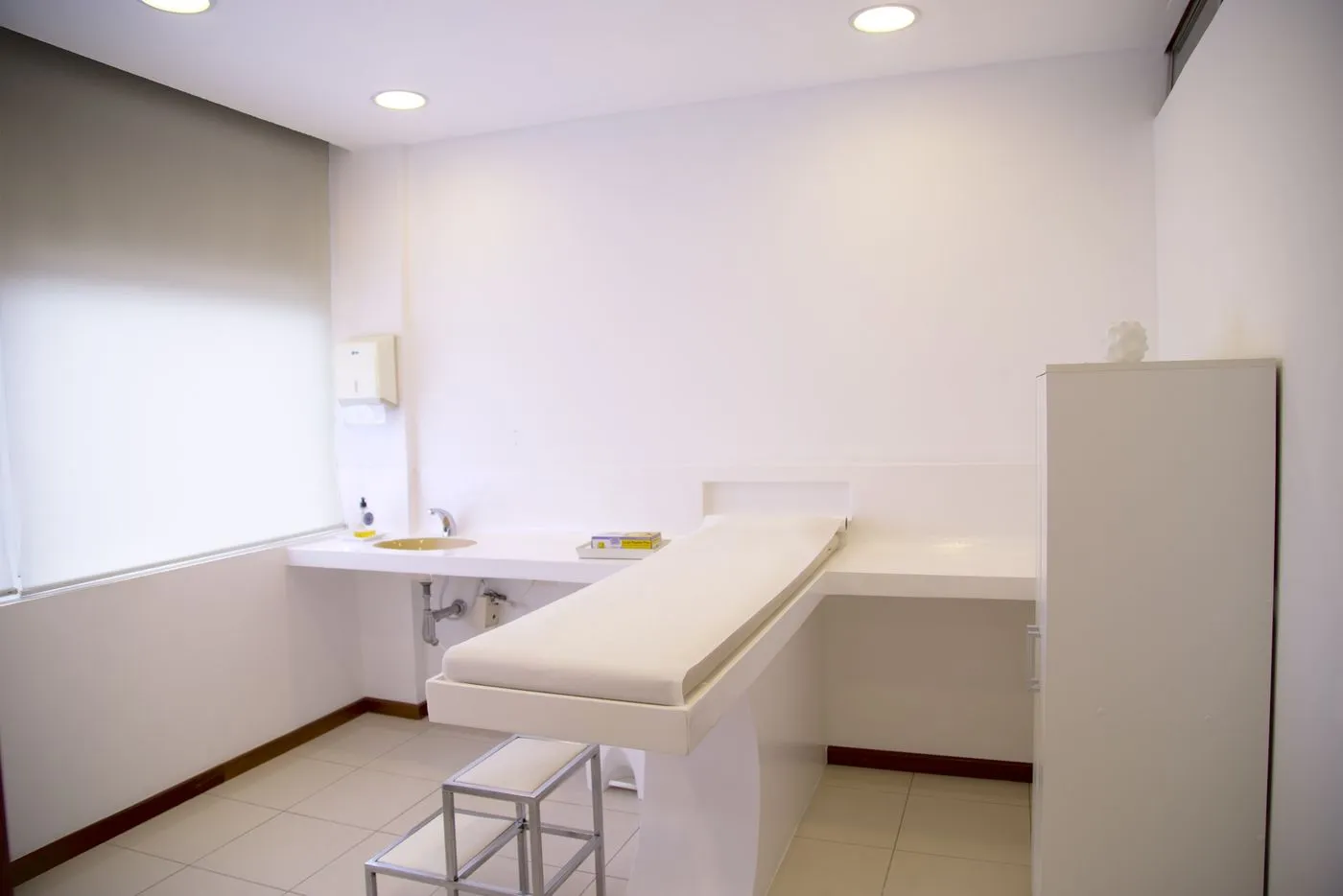 An empty, minimalist clinic examination room with a padded exam table, sink, and cabinet.