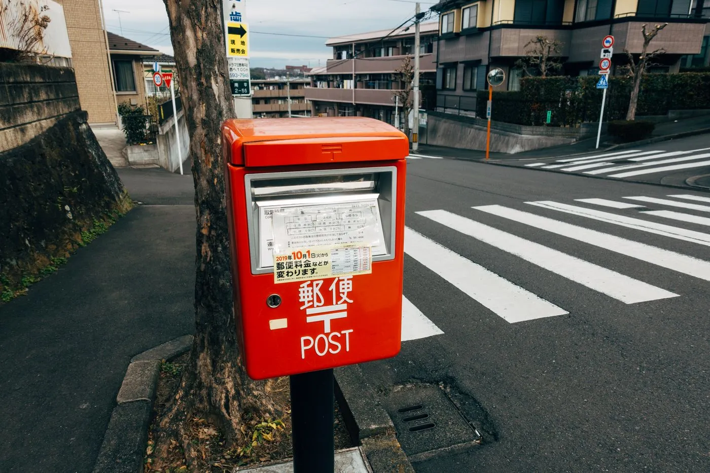A red Japanese postbox marked with post office signage, standing on a street corner near a crosswalk and residential buildings.