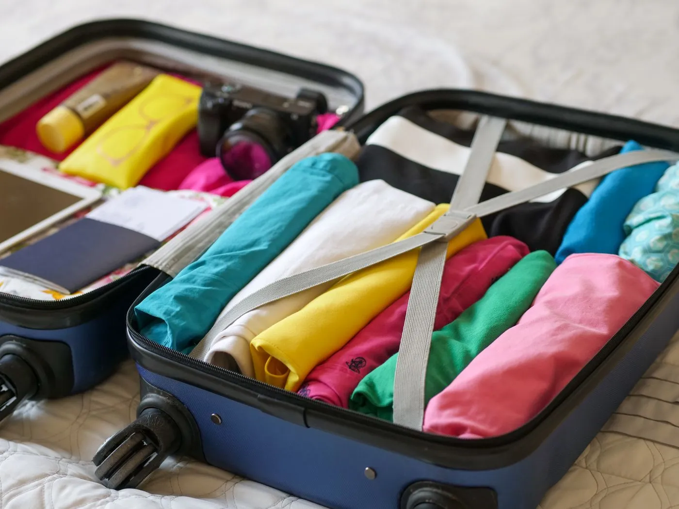 An open suitcase packed with rolled clothing in bright colors, secured with cross straps, with a passport tucked in the adjoining compartment.