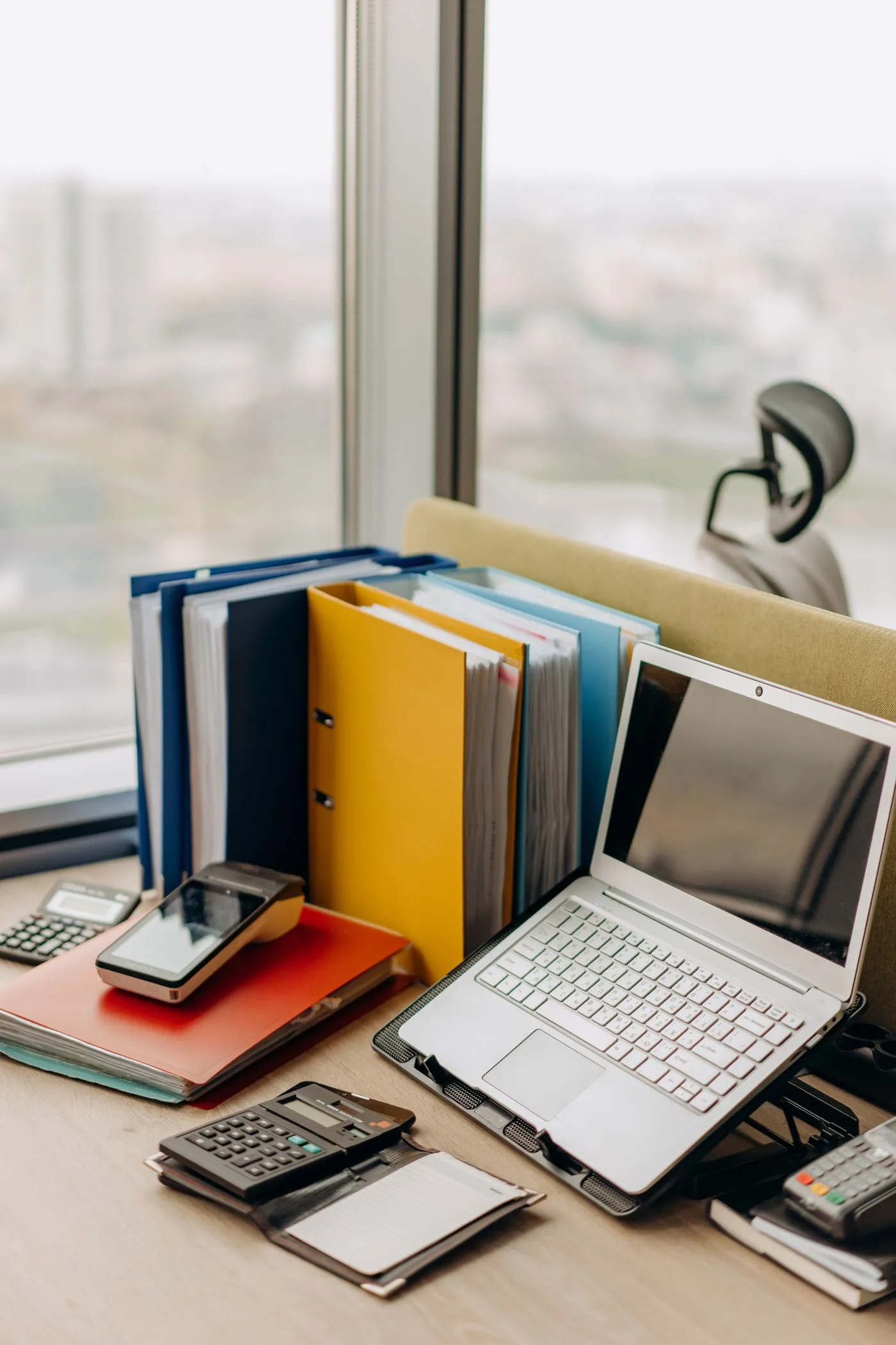 An office desk beside a window holding a row of ring binders, an open laptop, two calculators and a card payment terminal.