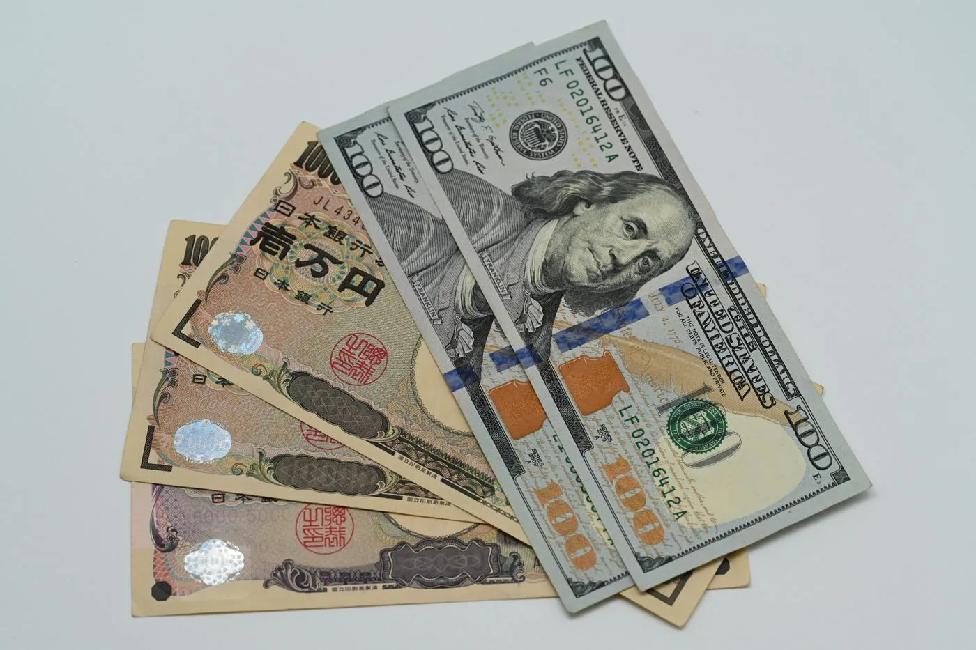 Japanese ten-thousand-yen notes fanned out underneath United States hundred-dollar bills on a pale surface.