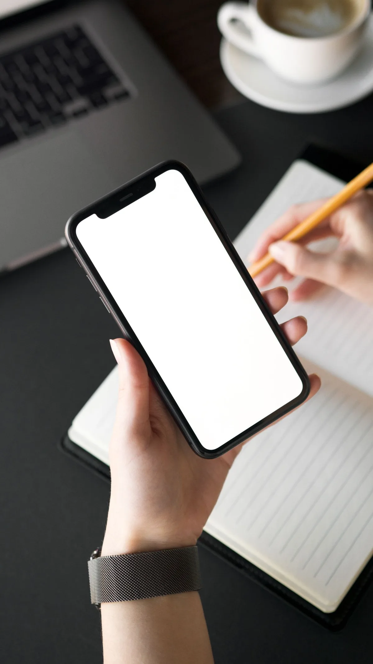 Hands holding a smartphone with a blank screen above an open notebook, with a laptop and a coffee cup on the desk behind.