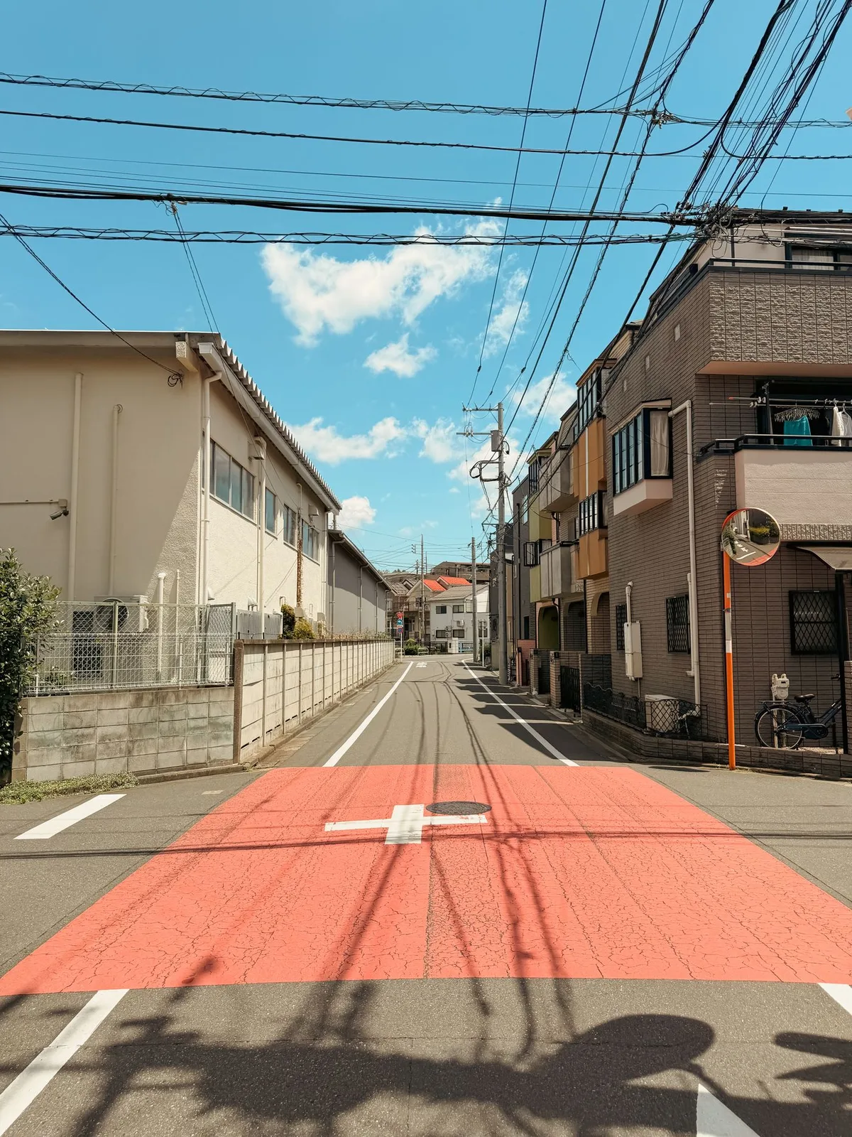 A quiet Tokyo residential street under a clear sky, with a red-painted crossing area and a convex traffic mirror.