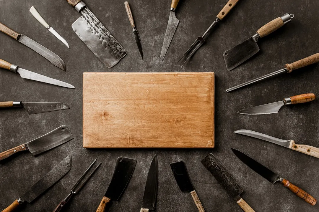 An overhead view of many kitchen knives and cleavers arranged around an empty wooden board.